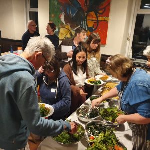 People of different ages and genders serving themselves food at a buffet in a crowded room. The room has two large windows and a large painted canvas with colourful abstract artwork on