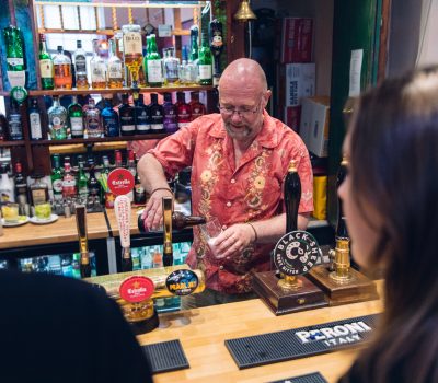 Behind a well-stocked bar, a man in a floral shirt pours a fruity cider for waiting customers