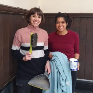 Two women stand smiling, one holding a dustpan and brush, and the other holding a paint can and sheet