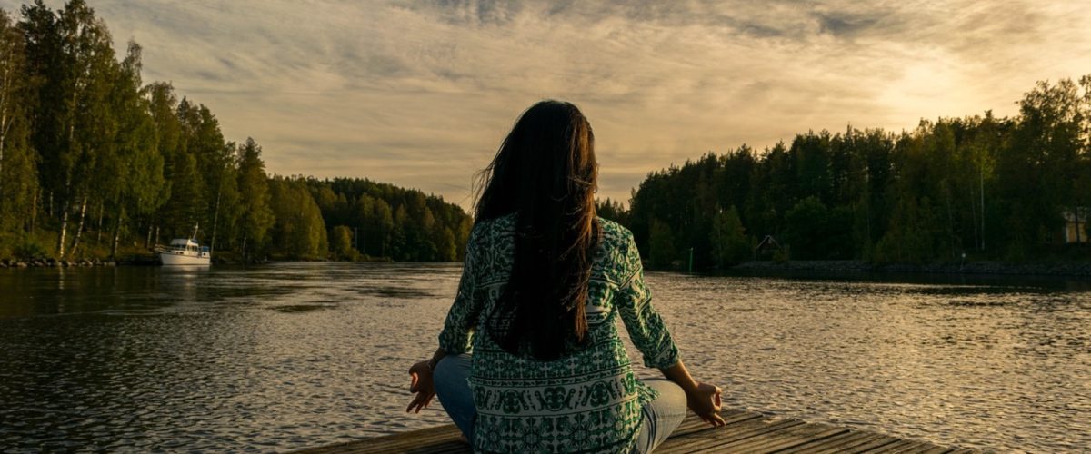 A woman sits meditating by a calm body of water