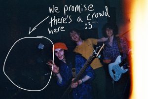 A band takes a selfie with an unseen audience that have been washed out by the images lack of exposure. It is annotated with "We promis there's a crowd"