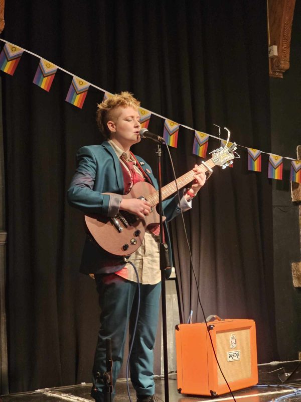 A musician with short hair and a suit plays a guitar surrounded by progress pride flags