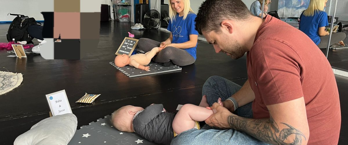 A dad sits cross-legged behind a baby on a small cushion.